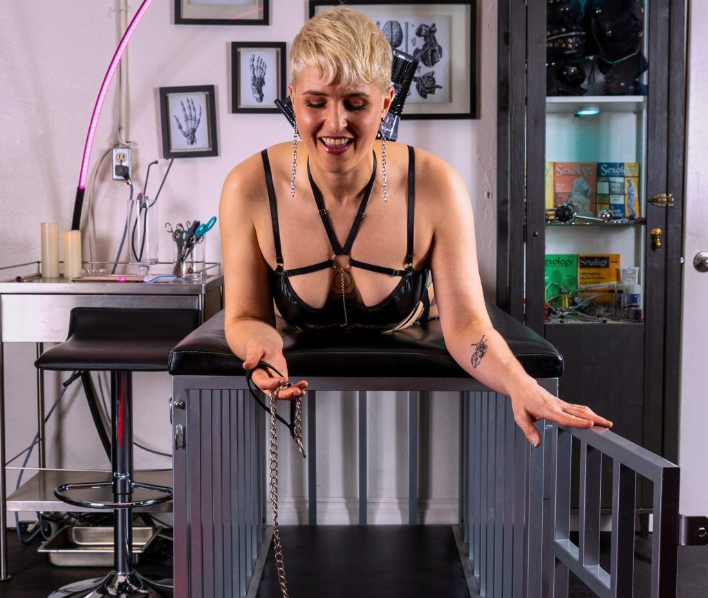 Queen Emma smiles and relaxes on top of a padded cage, holding the door open and dangling a leash.